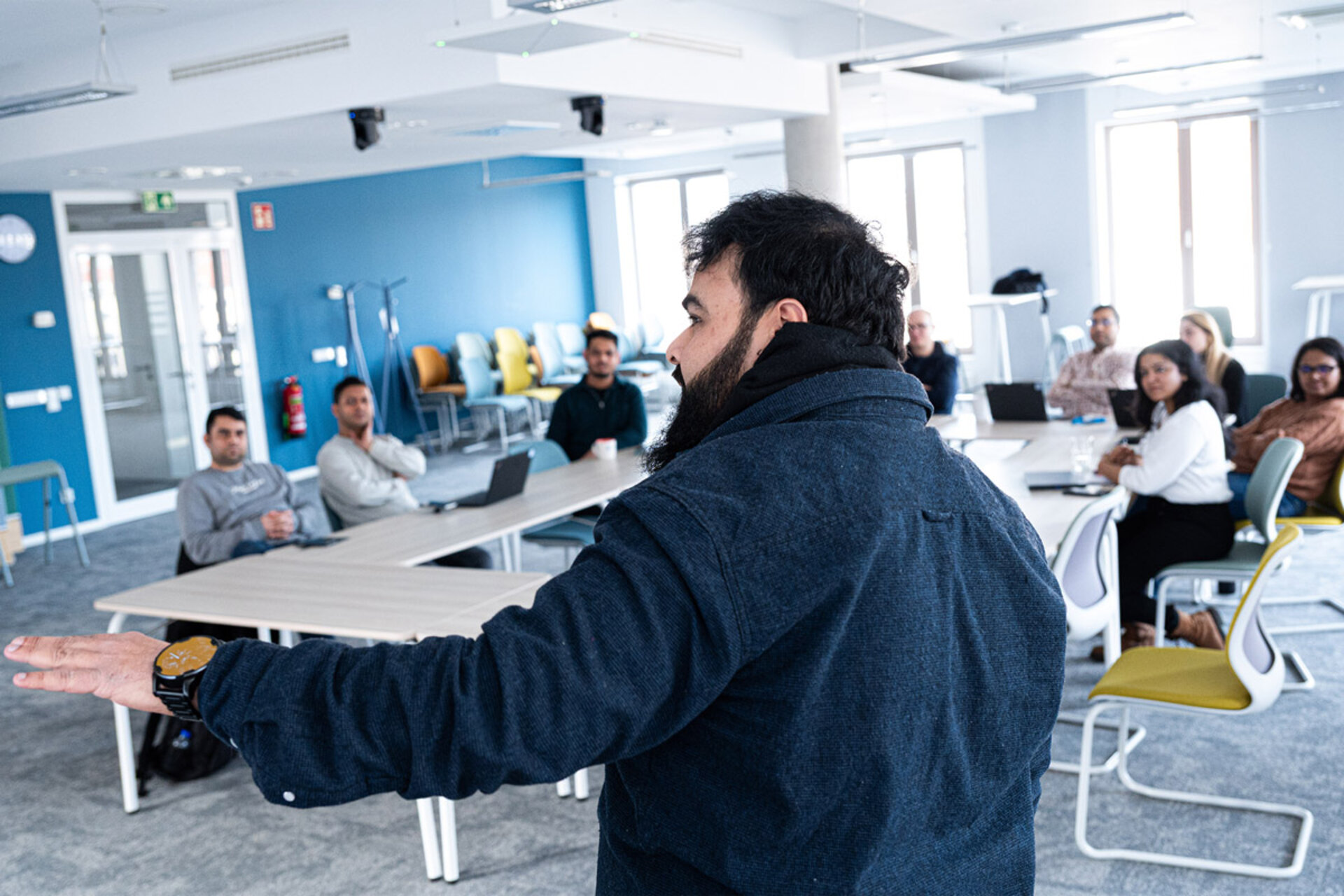 Person in dark jacket speaking to a group of seated people in a modern office space with blue walls and large windows