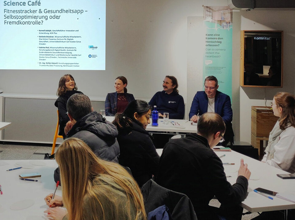In the foreground you see people sitting at 2 tables. Some of them are writing. Some others look at the 4 people who sit in the middlegound in a row talking. In the background you see written on a beamer slide: Science Café.
