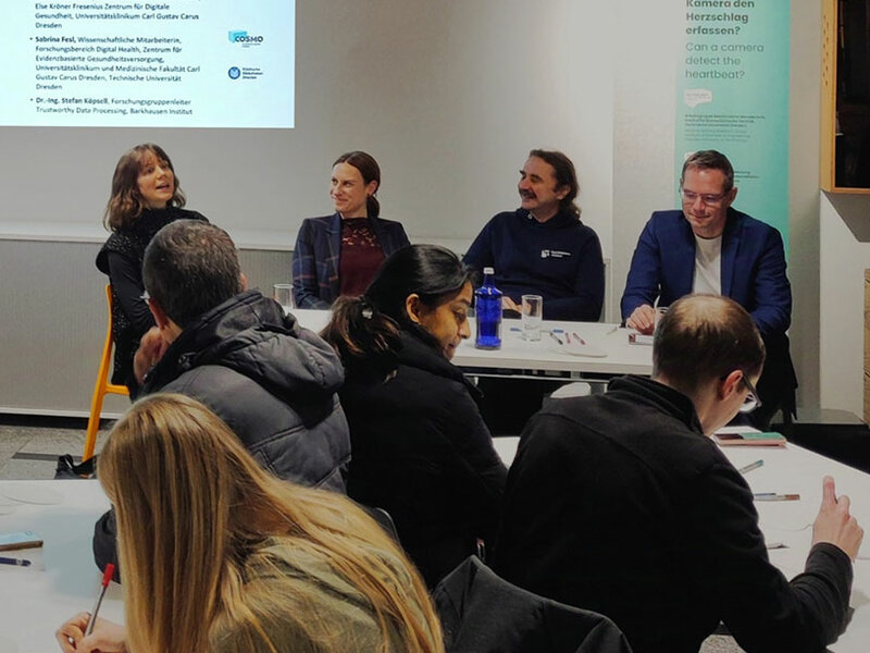 Im Vordergrund sieht man Menschen an 2 Tischen sitzen. Einige von ihnen schreiben. Einige andere schauen auf die 4 Personen, die in der Mitte in einer Reihe sitzen und sich unterhalten. Im Hintergrund sieht man eine Schrift auf einer Beamer- Leinwand: Science Café.