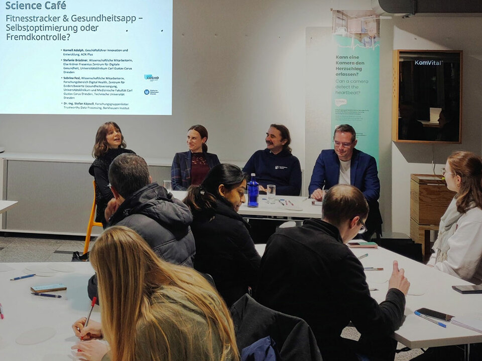 In the foreground you see people sitting at 2 tables. Some of them are writing. Some others look at the 4 people who sit in the middlegound in a row talking. In the background you see written on a beamer slide: Science Café.
