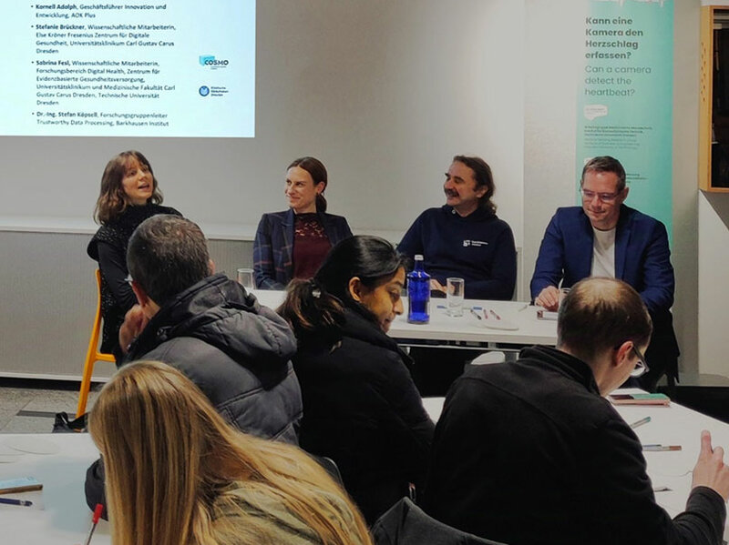 In the foreground you see people sitting at 2 tables. Some of them are writing. Some others look at the 4 people who sit in the middlegound in a row talking. In the background you see written on a beamer slide: Science Café.