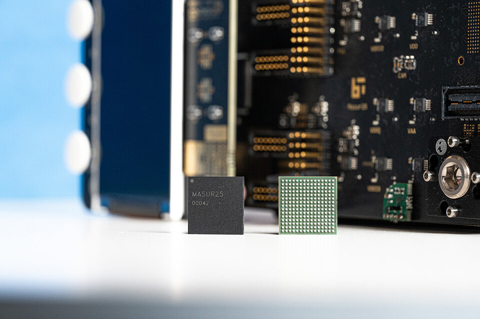 Das Gehäuse des Masur25 links mit der Aufschrift 'MASUR25 00042' und der Chip Masur25 rechts mit einer Matrix aus Kontaktflächen, im Hintergrund ein Evaluationsboard mit sichtbaren elektronischen Bauteilen.