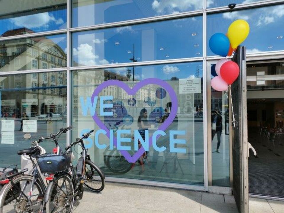 Glasfront eines Gebäudes mit der Aufschrift "We love science". Davor stehen Fahrräder und Luftballons hängen neben der Tür.
