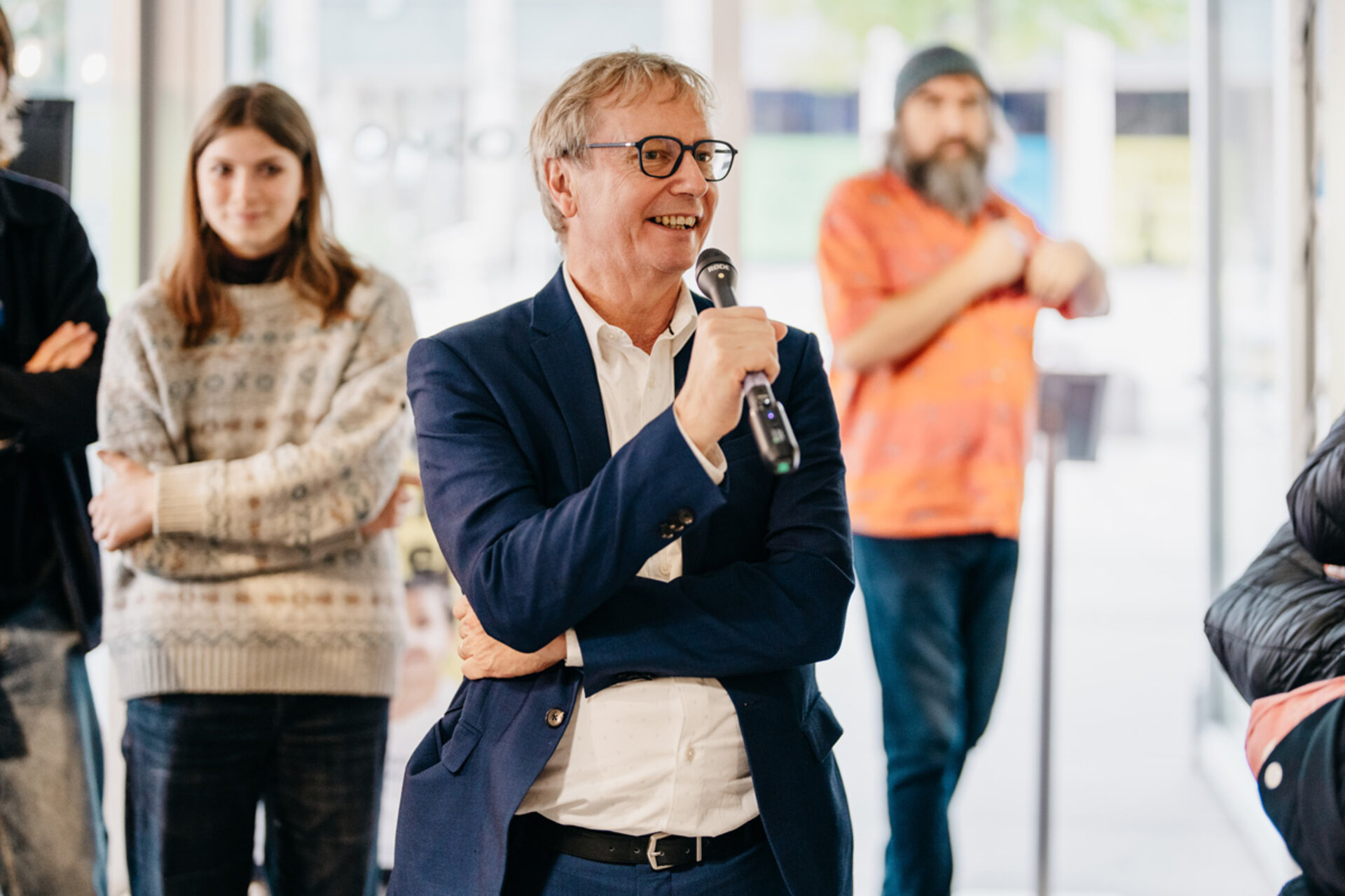 Man in blue blazer and white shirt (Prof. Gerhard Fettweis) holding microphone, with people in sweater and orange T-shirt in background at exhibition opening “Mensch, Roboter! – Robotik aus der Dresdner Wissenschaft.”