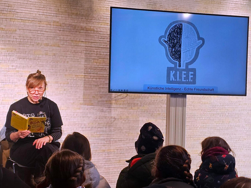 Frau sitzt auf einem Stuhl und liest aus einem gelben Buch vor vor Publikum, im Hintergrund Bildschirm mit Logo 'K.I.E.F' und Text 'Künstliche Intelligenz - Echte Freundschaft'