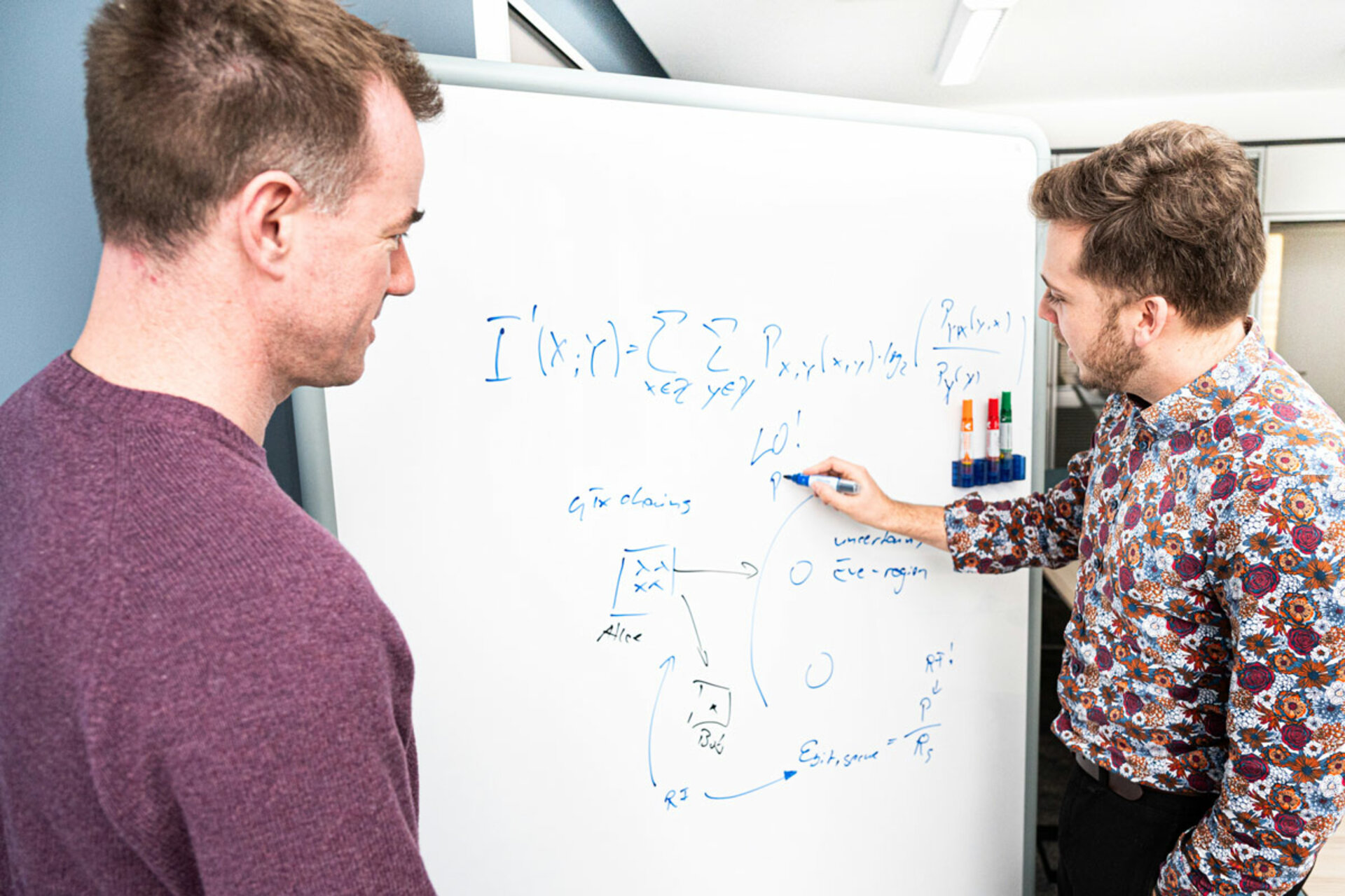 Zwei Männer stehen vor einem Whiteboard, auf dem mathematische Formeln und Diagramme mit blauer Schrift geschrieben sind.