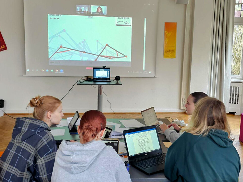 Vier Personen sitzen an einem Tisch mit Laptops und schauen auf eine Wandprojektion mit einem Diagramm und einer Videokonferenz.