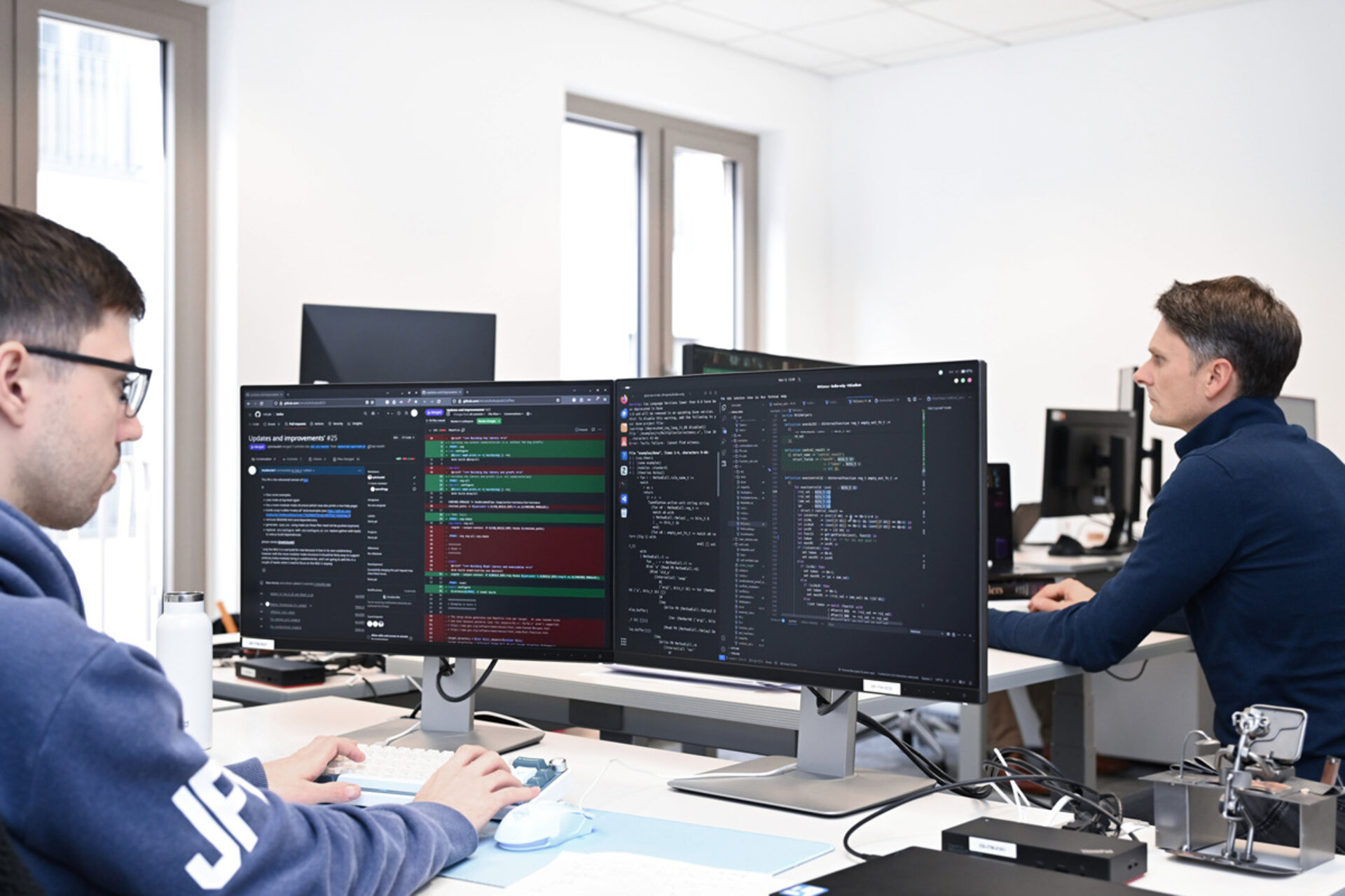 Zwei Personen sitzen an modernen Schreibtischen in einem hellen Büro. Beide arbeiten an Computern mit großen Monitoren. Auf den Bildschirmen sind Programmierumgebungen mit Quellcode und farblich hervorgehobenen Zeilen zu sehen. Die Arbeitsplätze sind aufgeräumt, und im Hintergrund stehen weitere Monitore und Büromöbel. Die Gesichter der Personen sind unkenntlich gemacht.