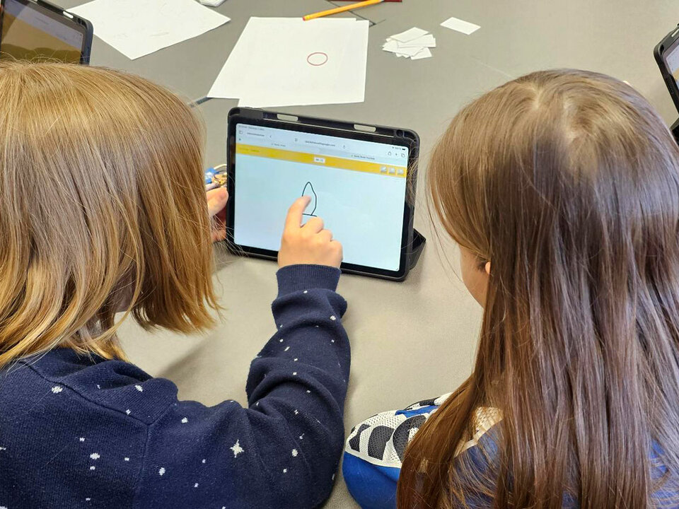 Zwei Kinder sitzen an einem Tisch und betrachten ein Tablet, auf dem eine einfache Zeichnung zu sehen ist.