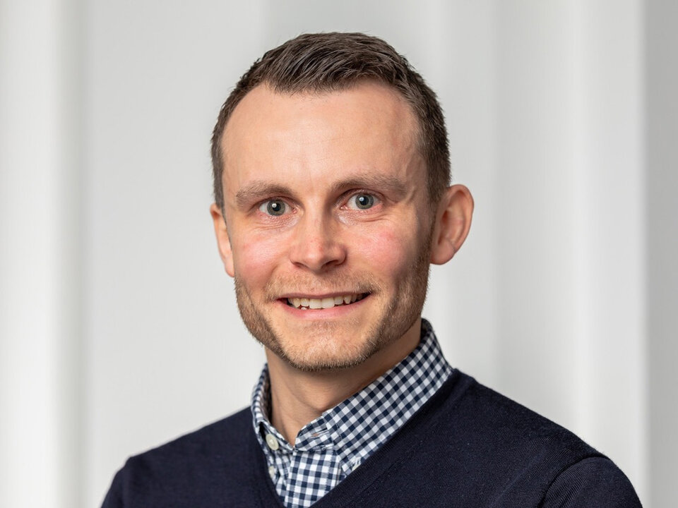 Mann mit kurzem Haar trägt ein kariertes Hemd unter einem dunklen Pullover vor hellem Hintergrund.