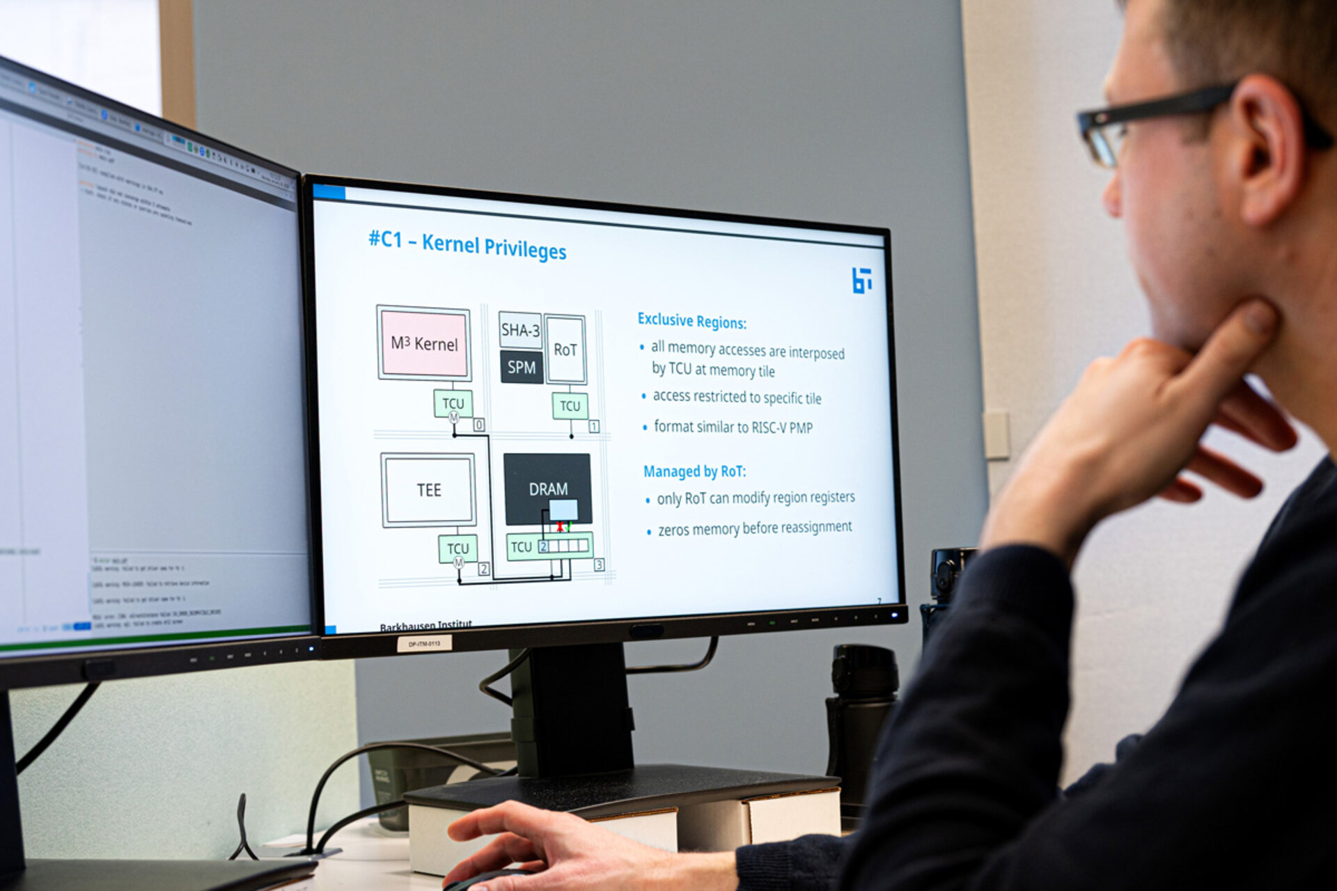 Person mit Brille sitzt vor zwei Monitoren, auf einem ist eine Präsentation zu Kernel-Privilegien mit Diagrammen und Text zu sehen