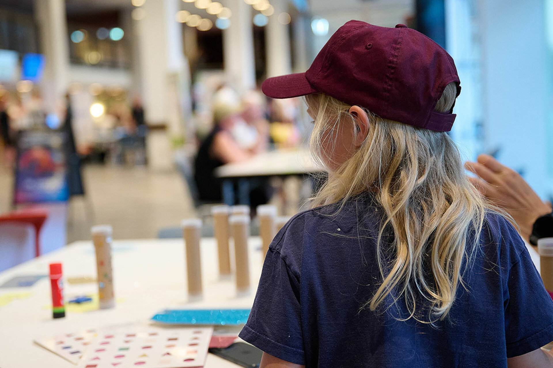 Kind mit langem blondem Haar und roter Kappe sitzt an einem Basteltisch in einer Halle mit unscharfem Hintergrund.