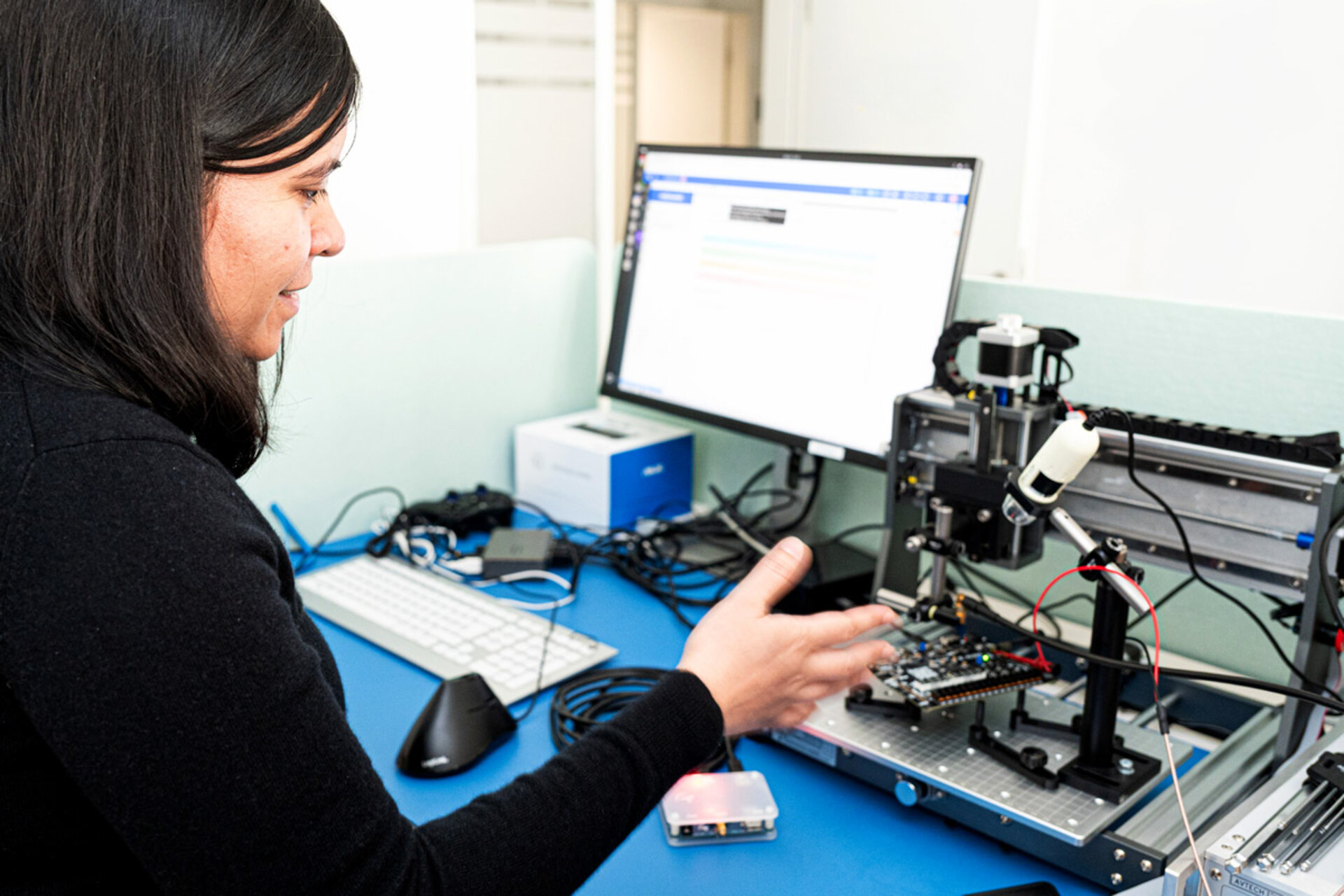 Person sitting at a desk with a computer monitor, keyboard, mouse, and an electronic device with wiring and mechanical parts