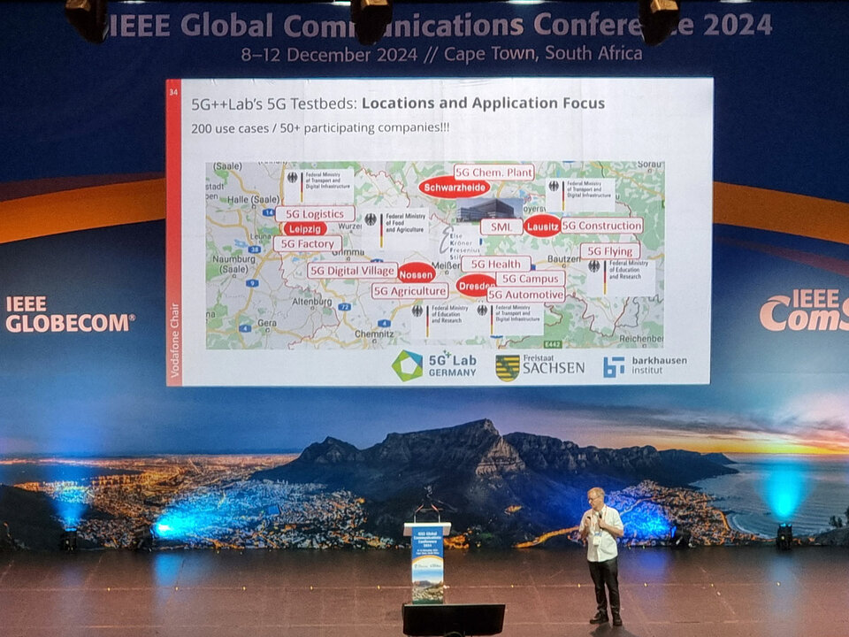 Gerhard Fettweis speaks at IEEE GLOBECOM 2024 in Cape Town about the 5G++Lab testbeds. The presentation slide shows various 5G applications and locations in Saxony. The scenic backdrop of Cape Town is visible in the background.