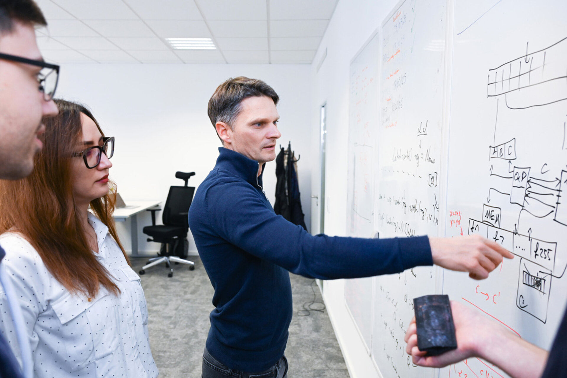 Drei Personen stehen vor einer Wand mit Diagrammen und Formeln, eine Person zeigt mit dem Finger darauf