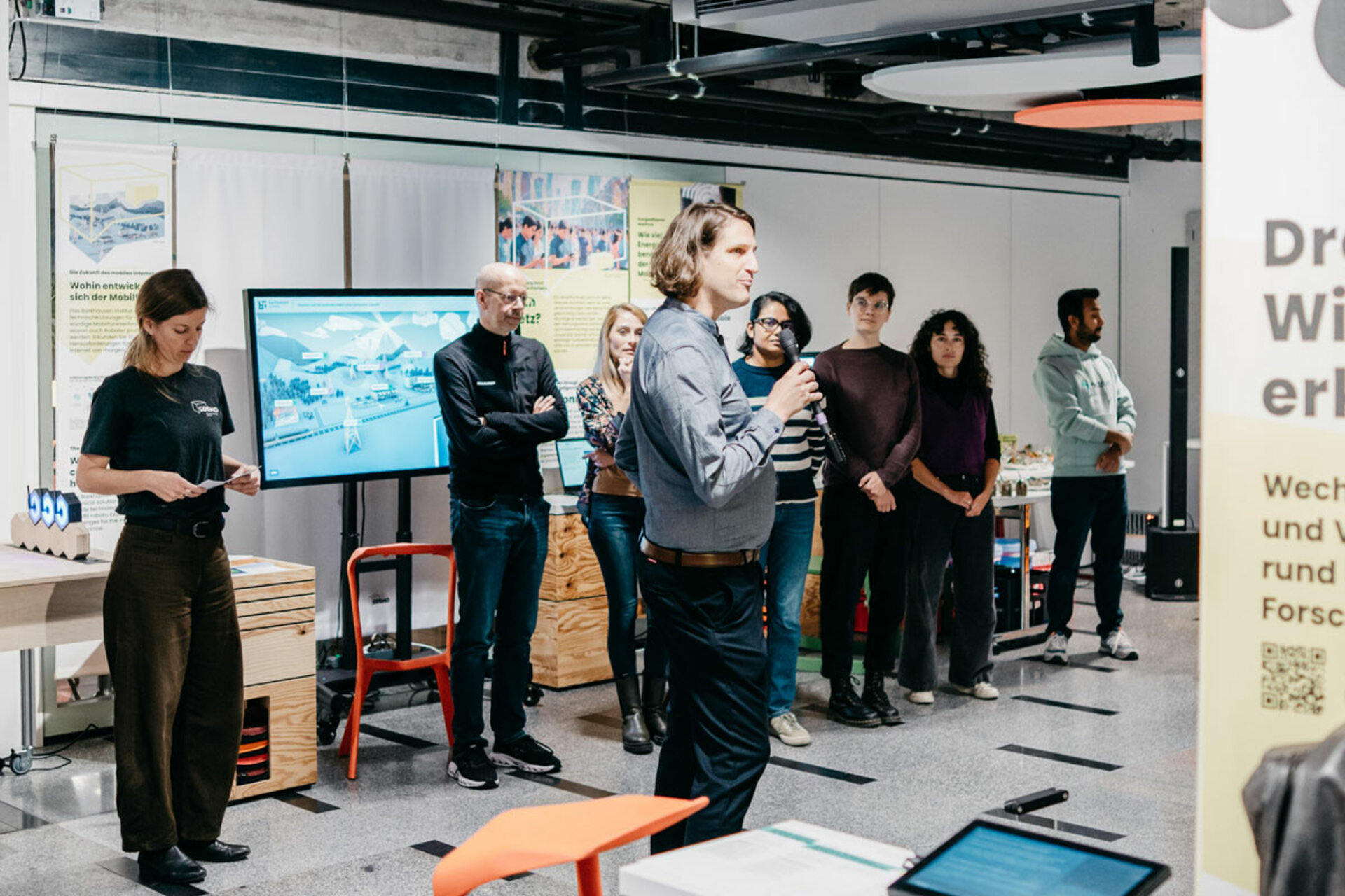 Person (Dr. Konstantin Klamka) holding microphone speaking to a group at the opening of the exhibition 'Mensch, Roboter! – Robotik aus der Dresdner Wissenschaft' in a modern interior with monitor and display panels.