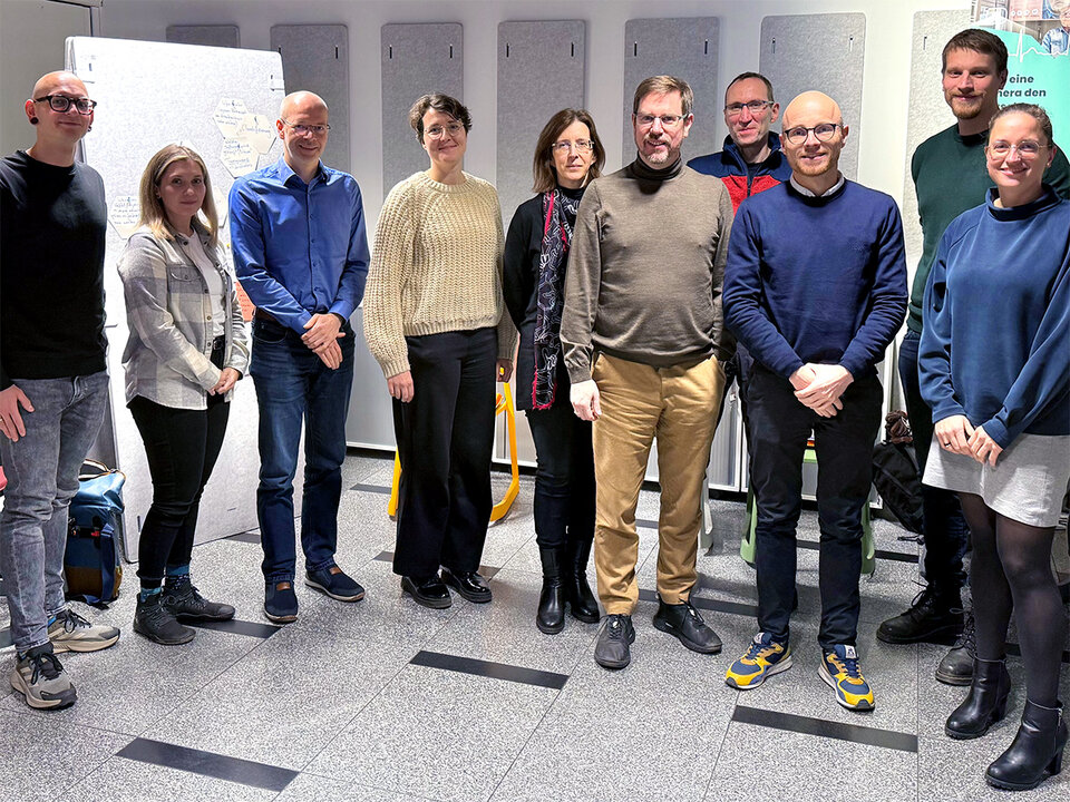 A group of ten people stands together in a bright room for a group photo. In the background, gray acoustic panels can be seen on the wall, and there is a flip chart on the left. The people are dressed differently and smiling at the camera.