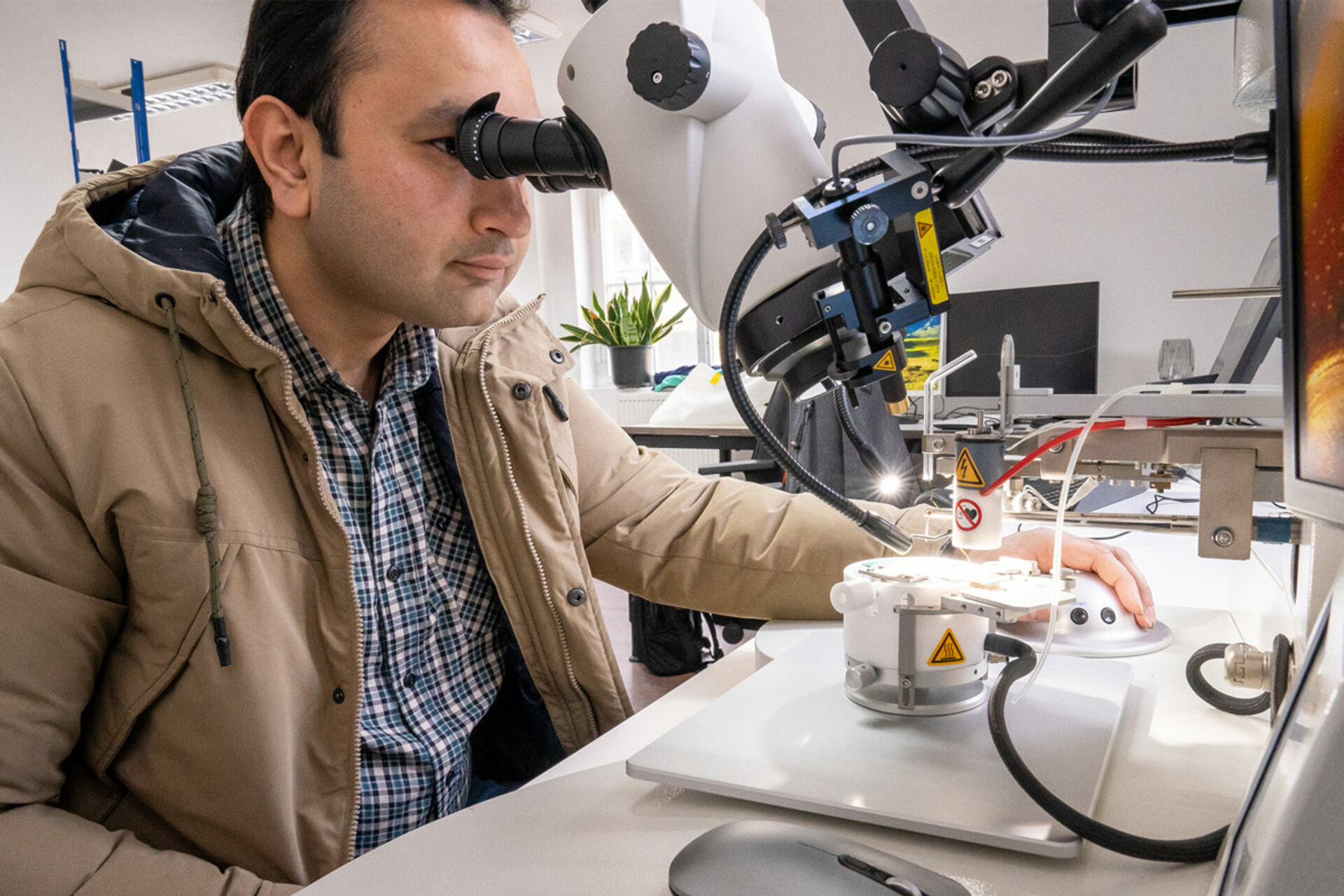 Person in a brown jacket and checkered shirt sitting at a table operating a robotic arm with a white base and control panel