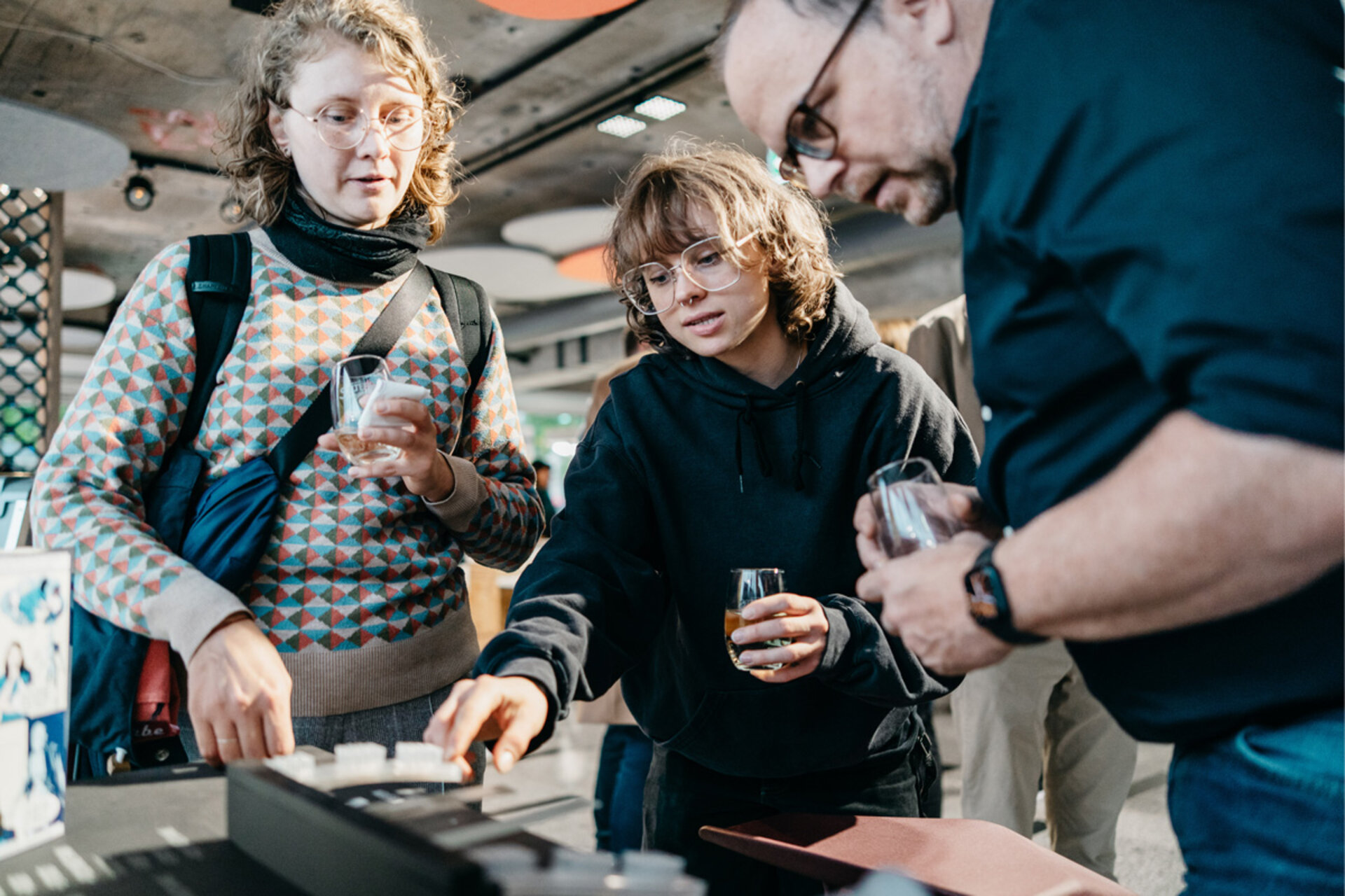 Three people stand around a table with soft material for robotic hands, one holding a glass, another touching the exhibit.