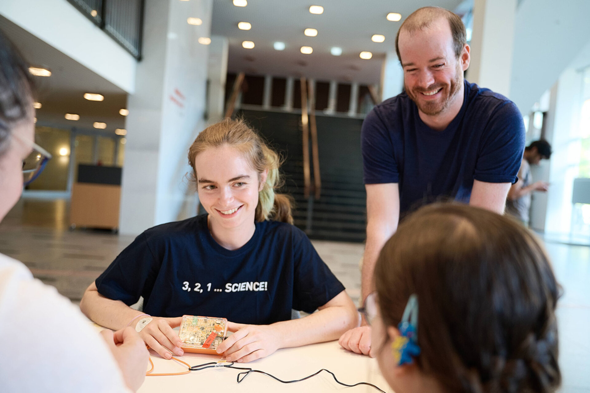 Junge Frau mit T-Shirt '3, 2, 1 ... SCIENCE!' sitzt an einem Tisch und zeigt ein elektronisches Gerät, während ein lächelnder Mann und zwei weitere Personen ihr zuhören.