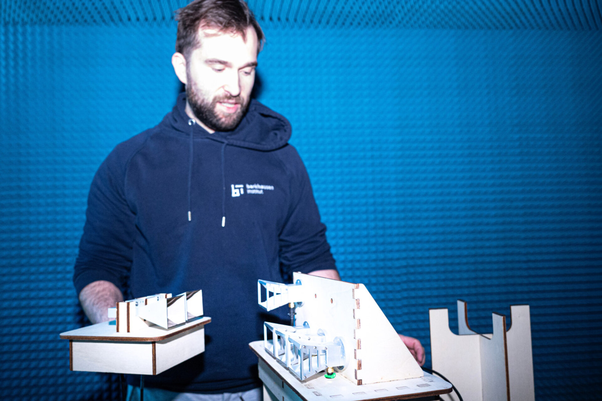 Person mit Kapuzenpullover hält zwei Modelle aus Holz oder Karton vor blauem Hintergrund