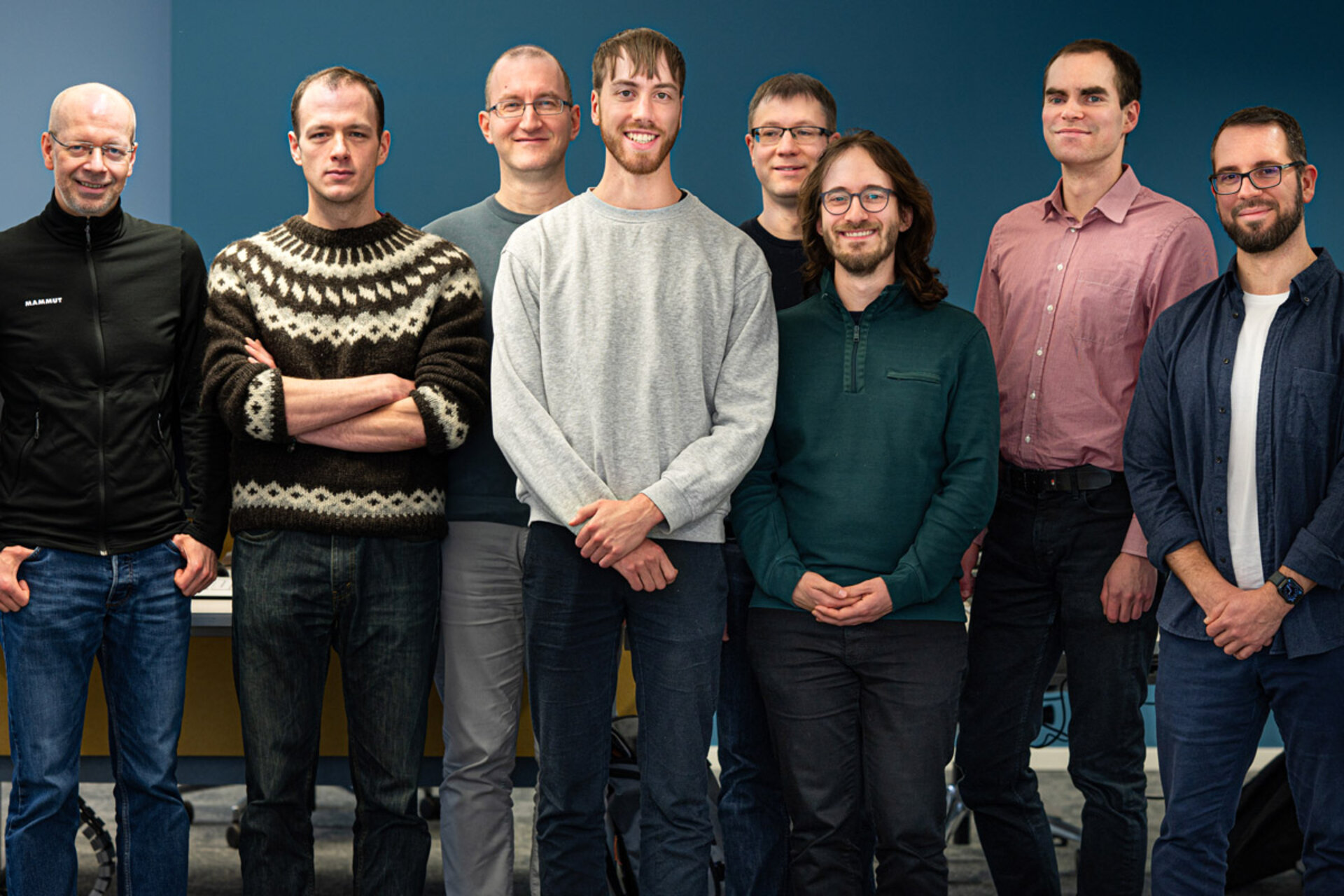 Gruppe von acht Personen steht nebeneinander vor einem dunkelblauen Hintergrund.
