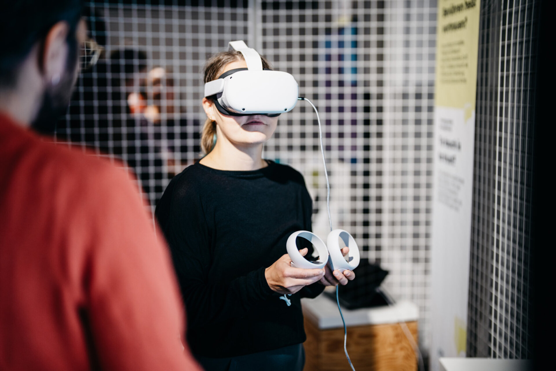 Person wearing a VR headset holding two white VR controllers, standing in front of a grid wall at an exhibition on robotics from Dresden science.