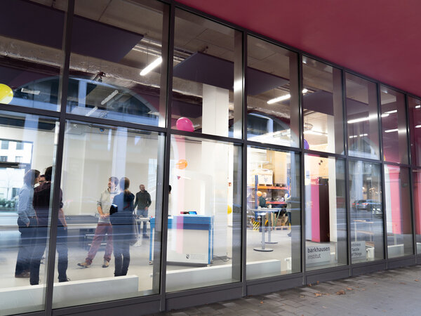 The picture shows the glass facade of a building through which you can see into a room. In this room, a few people are standing together in small groups and talking. There are also some pieces of furniture and a few colorful balloons are attached to the ceiling. The room looks modern and bright. There is a sign on the outside of the windows that reads “Barkhausen Institute”. The overall impression is that of a modern, open and inviting facility, possibly a research or work area.
