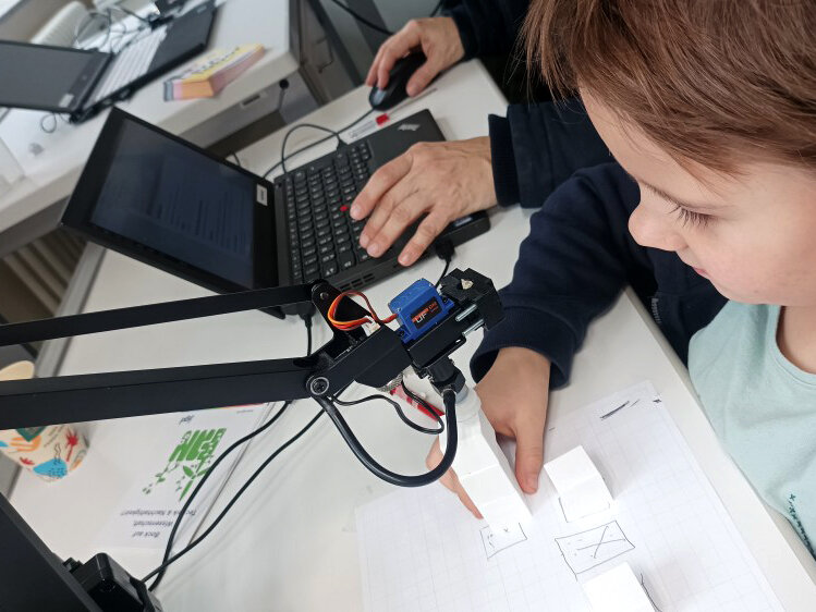 A child and a man sitting at a table, the man is working on a laptop and the child is interacting with a robot arm