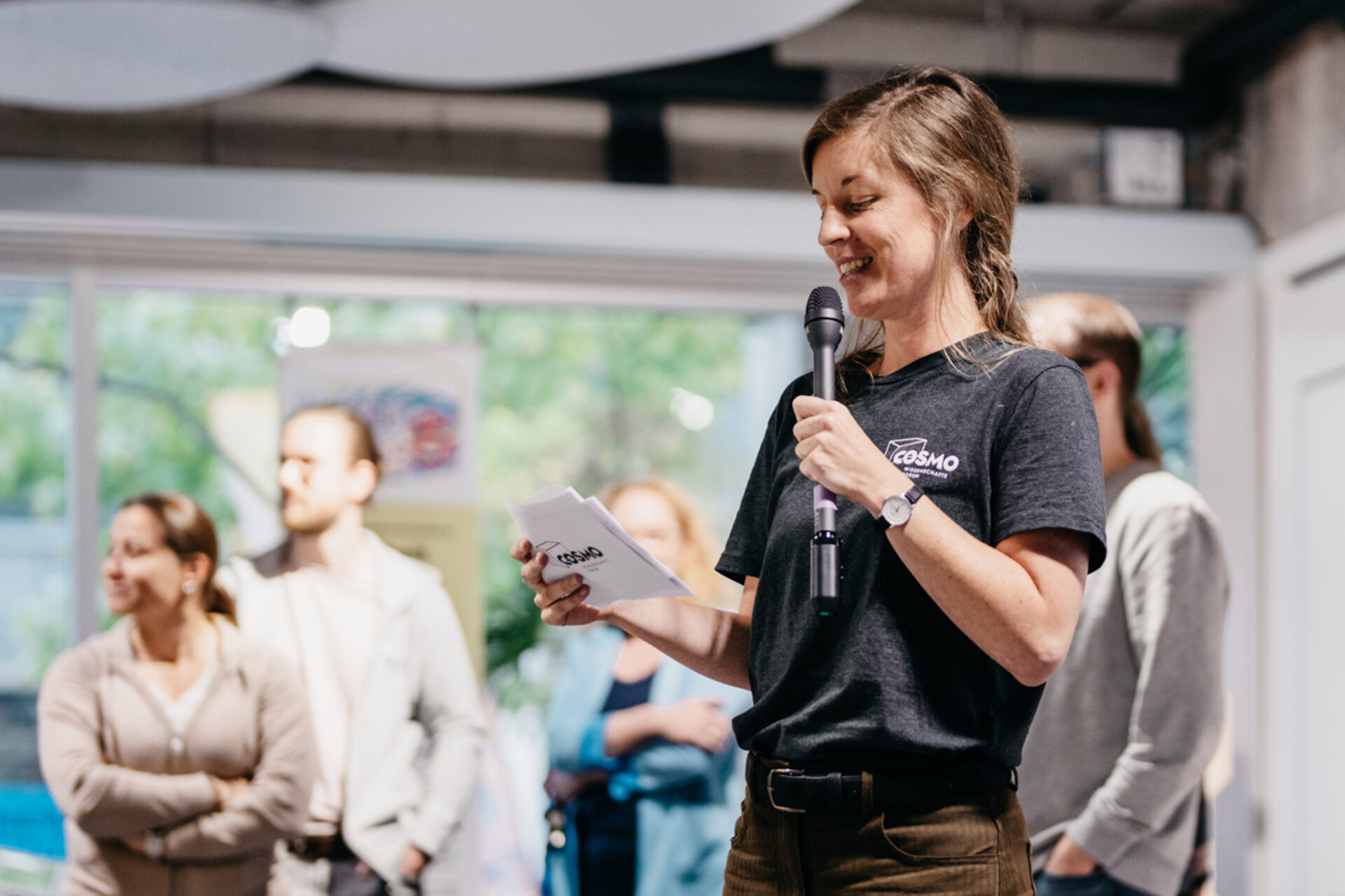 Woman with braid (Dr. Tiphaine Cattiau) wearing black 'COSMO' T-shirt holding microphone and notes at the opening of the exhibition 'Mensch, Roboter! – Robotik aus der Dresdner Wissenschaft'.