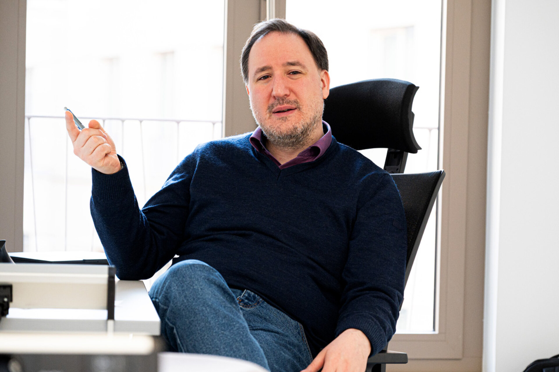 Man sitting in an office chair with one leg crossed over the other holding a pen in one hand