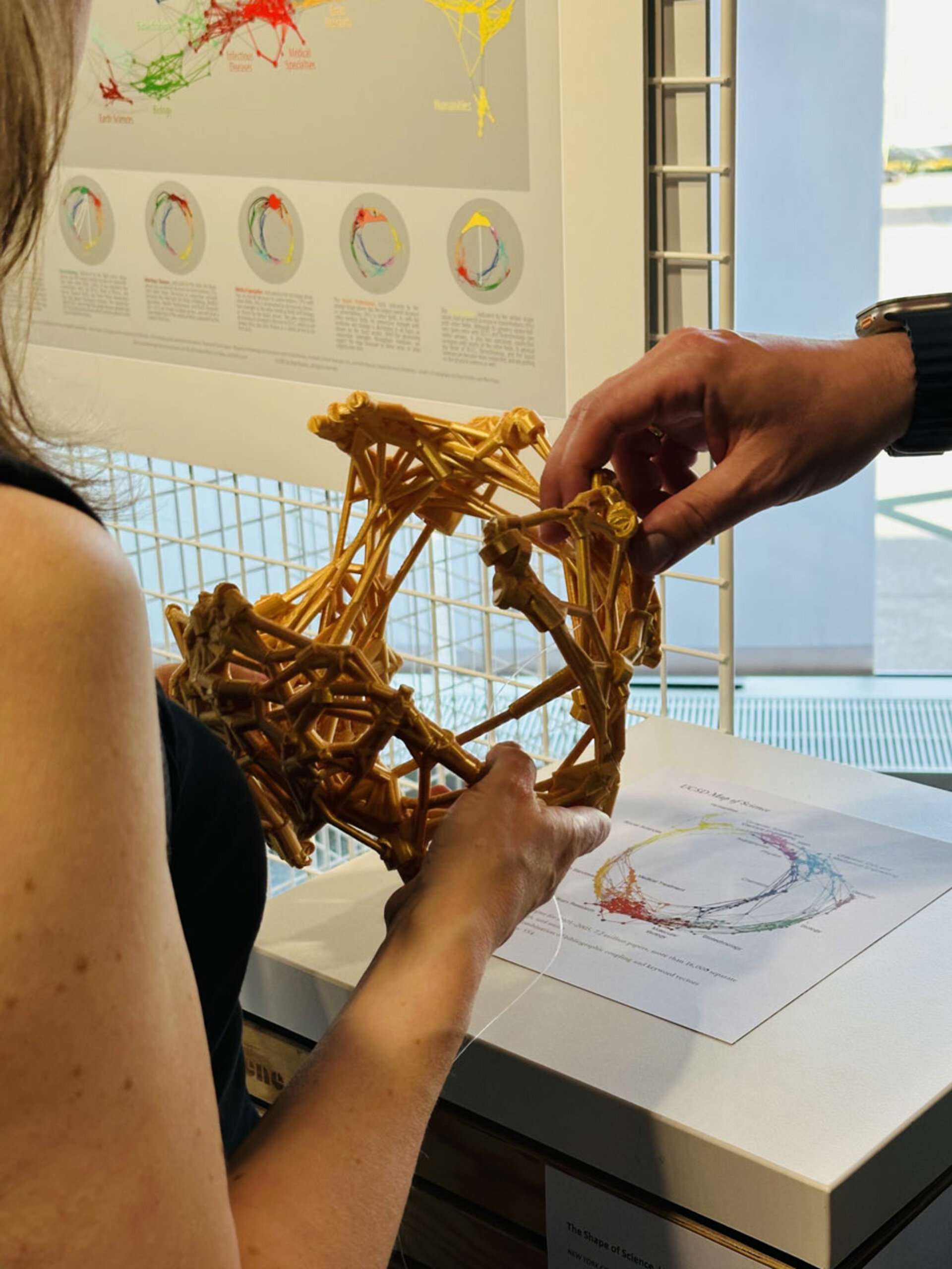 Eine Person hält eine komplexe, geometrische Skulptur, während eine andere Hand sie justiert. Dahinter ist ein wissenschaftliches Poster mit bunten Grafiken und Diagrammen zu sehen.