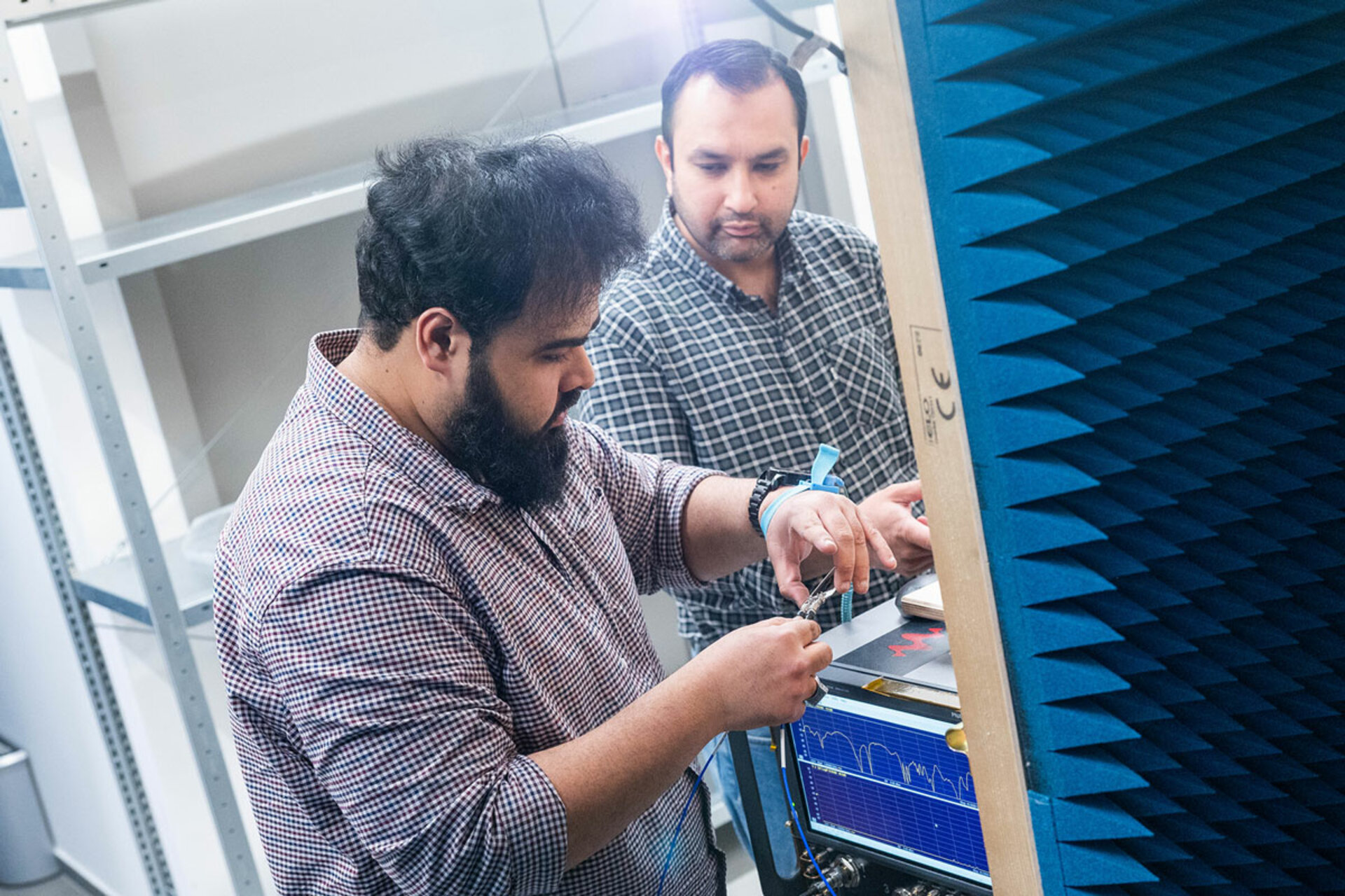 Two men working with electronic equipment in a room with blue acoustic foam panels