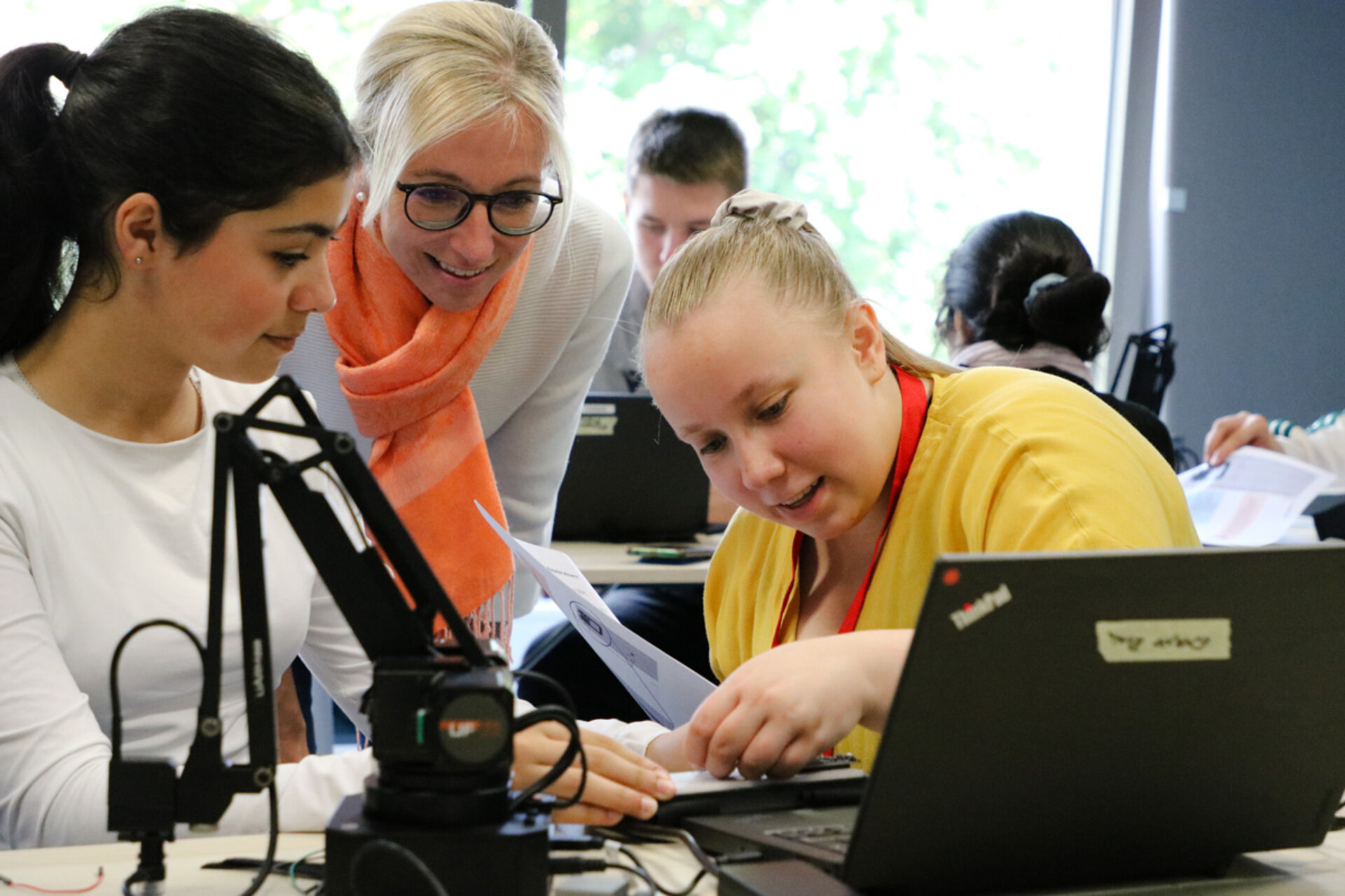 Three women collaborate in a classroom setting. One woman points at a laptop screen, while the others observe, creating a focused and engaged atmosphere. In the foreground, you can see a robot arm.