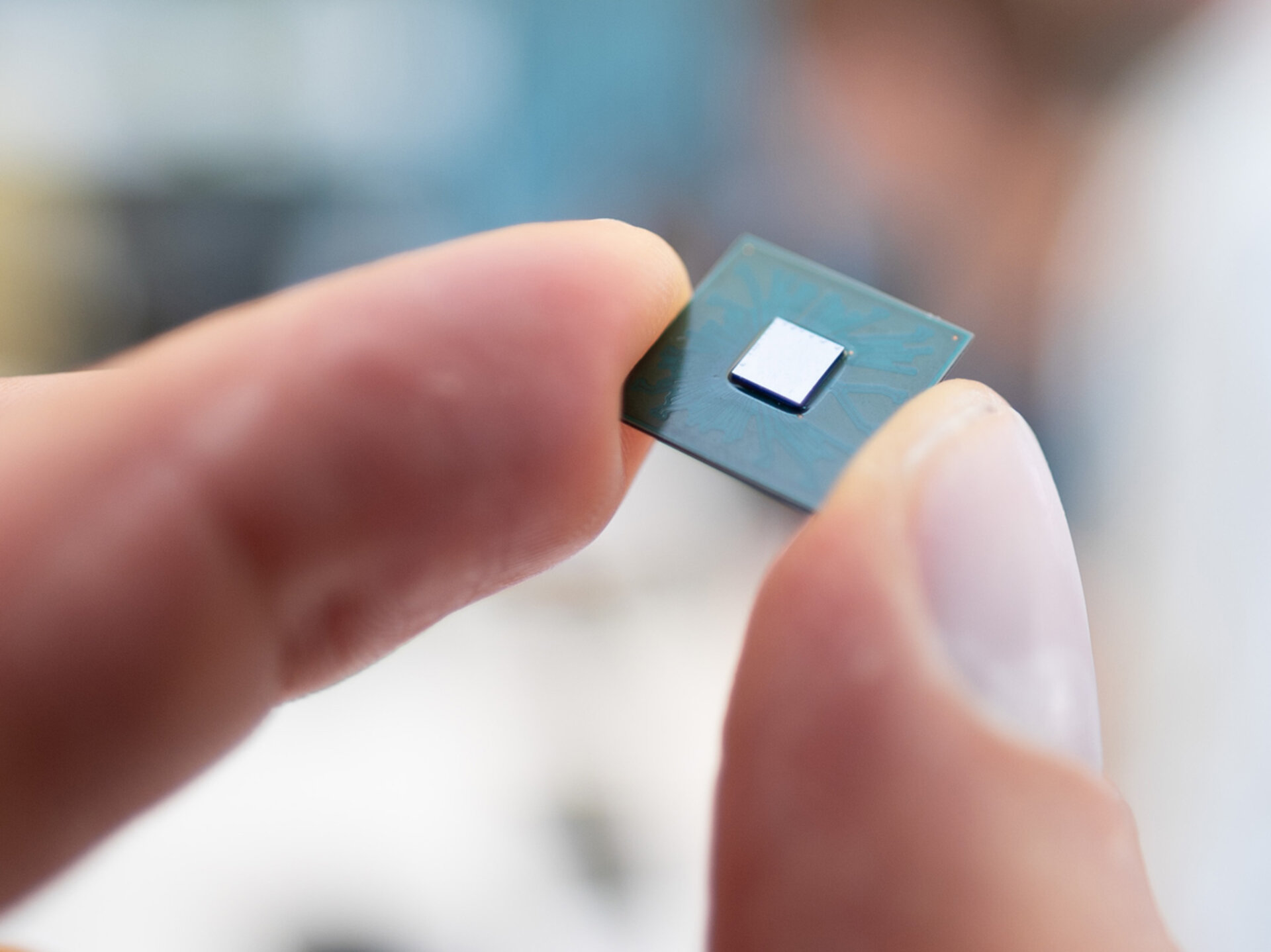 Eine Hand hält zwischen Daumen und Zeigefinger einen kleinen, quadratischen Chip mit einem weißen quadratischen Element in der Mitte. Der Hintergrund ist unscharf, sodass die Hand und der Chip im Vordergrund deutlich hervorgehoben sind.
