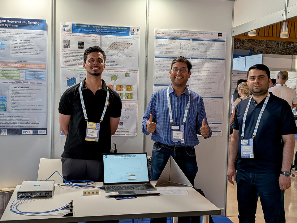 Three men standing behind a table with a laptop and electronic equipment in front of scientific posters