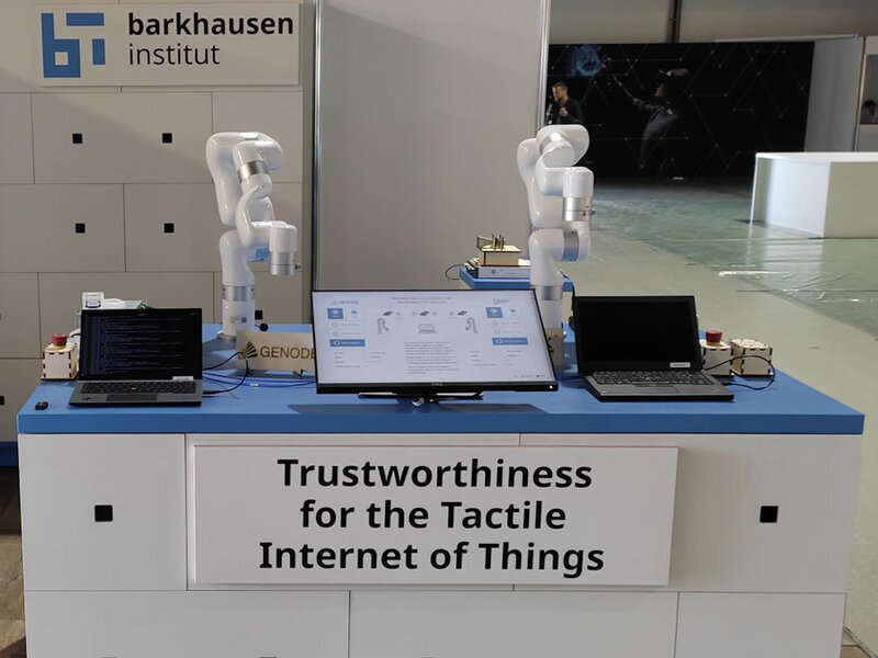 Zwei weiße Roboterarme auf einem blauen Tisch mit zwei Laptops, Schild mit Text 'Trustworthiness for the Tactile Internet of Things', Hintergrund mit Logo des Barkhausen Instituts.