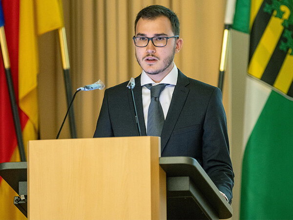 Andrey Ruzhanskiy holding his speech behind a standing desk. He is talking into a microphone. Behind him there are the flags of Germany and Saxony.