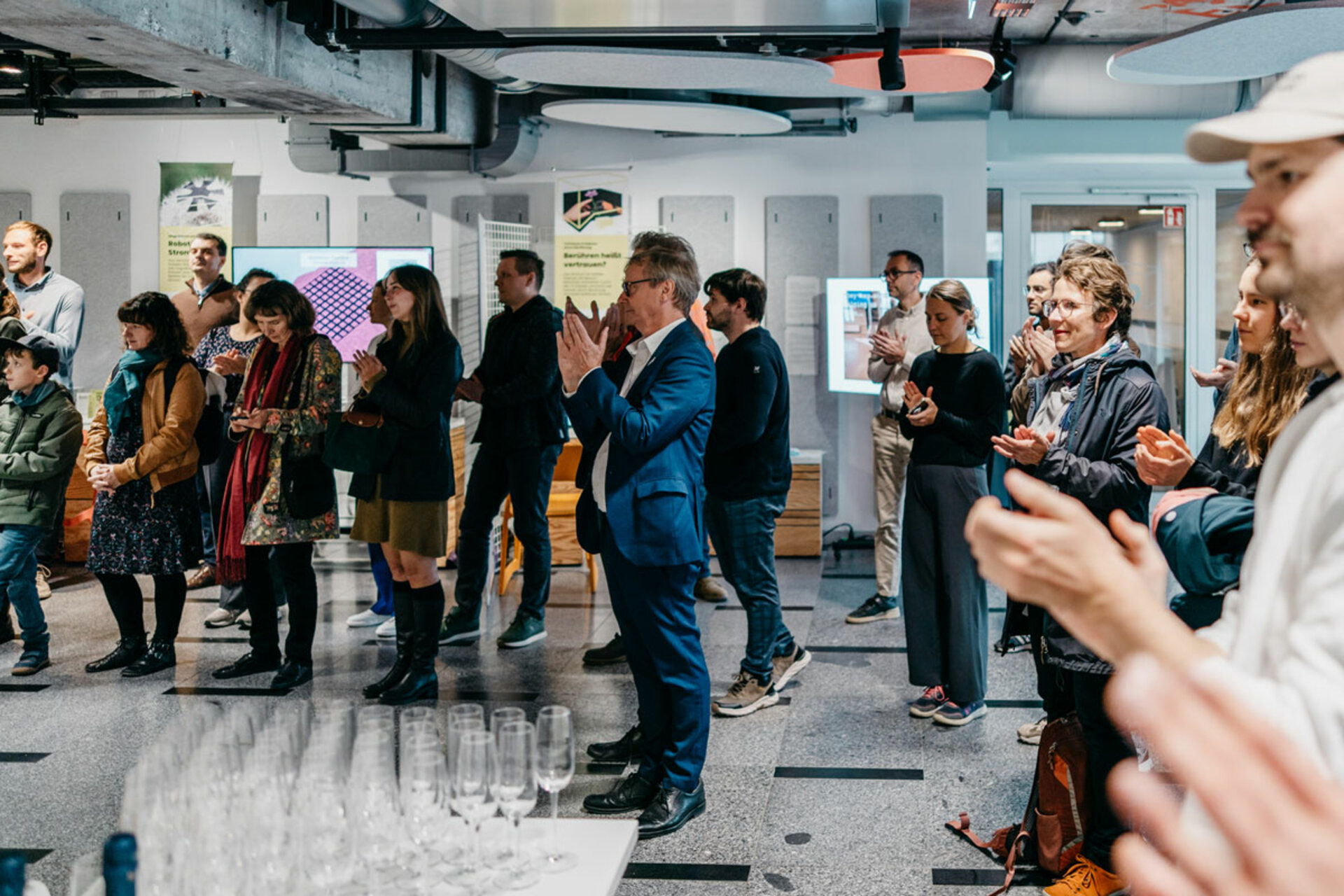 Several people standing in an exhibition space applauding. Empty champagne glasses on a table in the foreground. Exhibition panels and ceiling lights visible in the background.