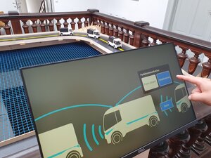 Auf dem Bildschirm sieht man Autos, welche über Funk kommunizieren. Hinter dem Bildschirm ist eine Modell-Autobahn mit eben diesem Konvoi zu sehen.