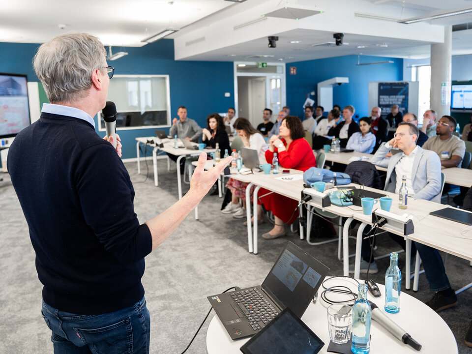 You can see an over-the-shoulder shot of the audience. The audience sits behind rows of tables and listens attentively to Prof. Fettweis' speech.