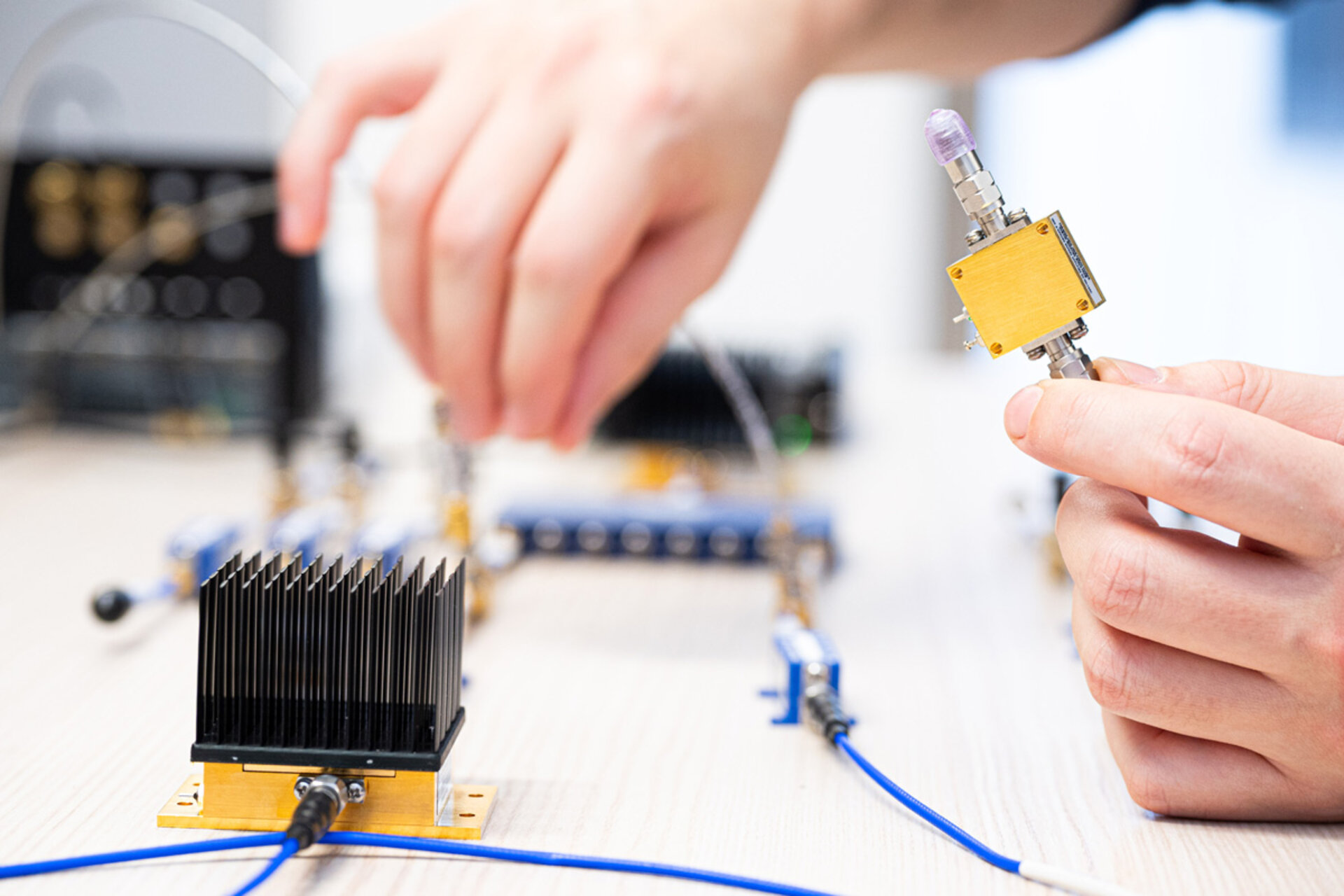 Hand hält ein kleines elektronisches Bauteil mit Kabelanschluss über einem Tisch mit weiteren elektronischen Komponenten und Kabeln