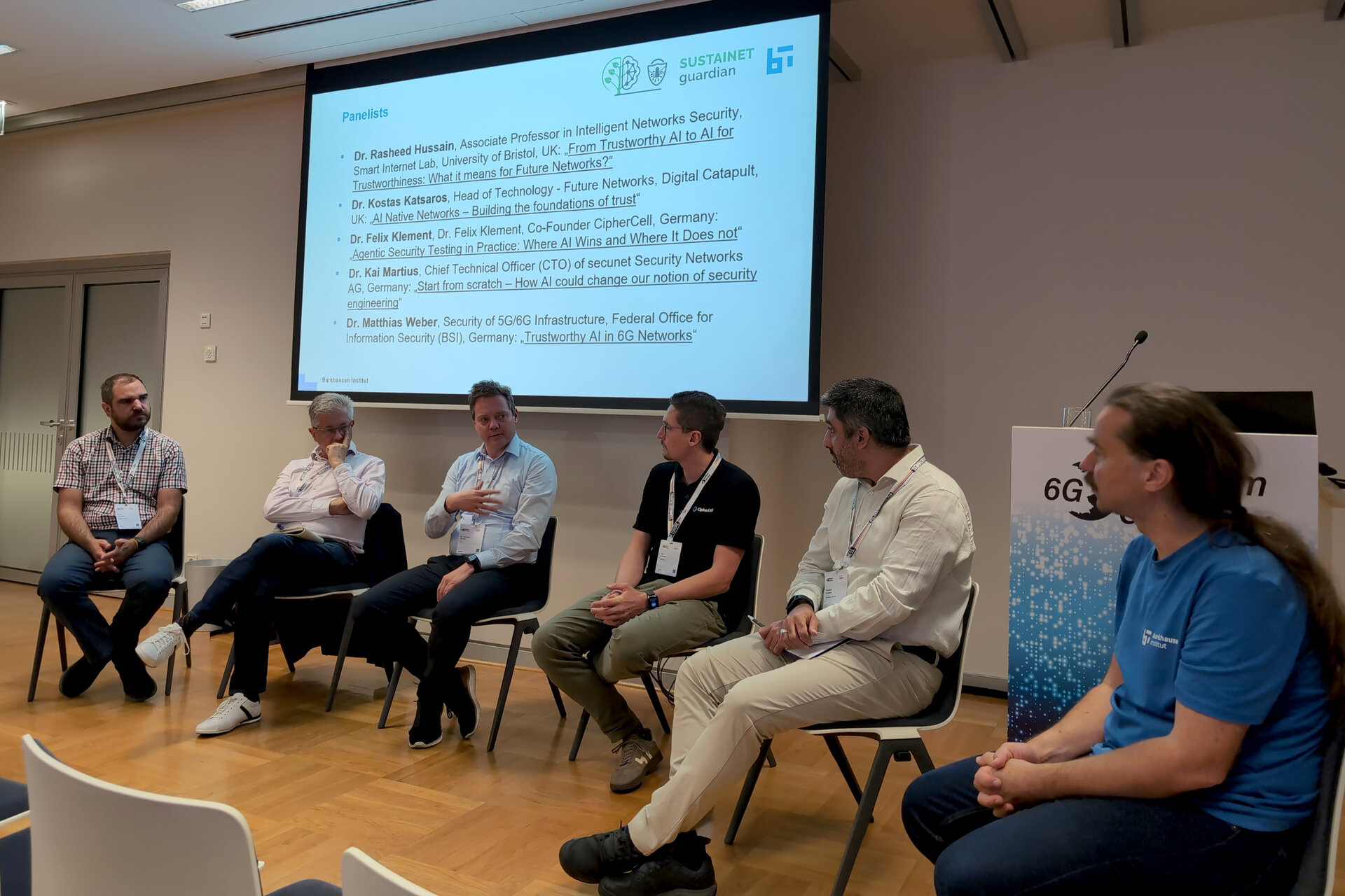Five men seated in a panel discussion in a conference room with a presentation slide behind them and a 6G conference banner visible