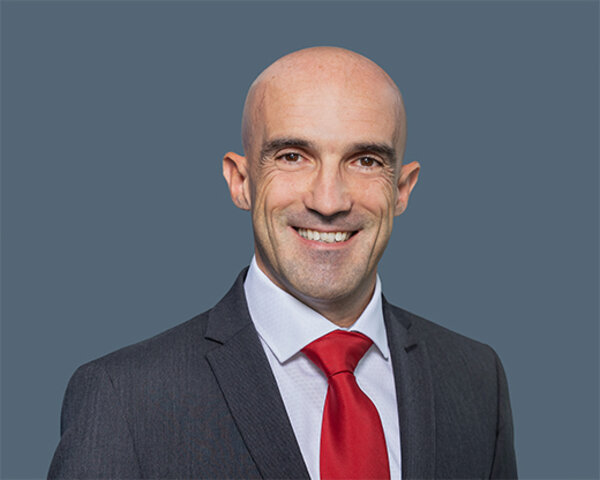 Mann mit Glatze trägt dunklen Anzug, weißes Hemd und rote Krawatte vor einfarbigem Hintergrund