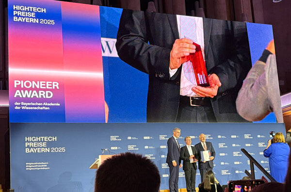 The three men stand on the stage for a group photo. The large screen behind the stage has zoomed in on the red award and shows it close up.