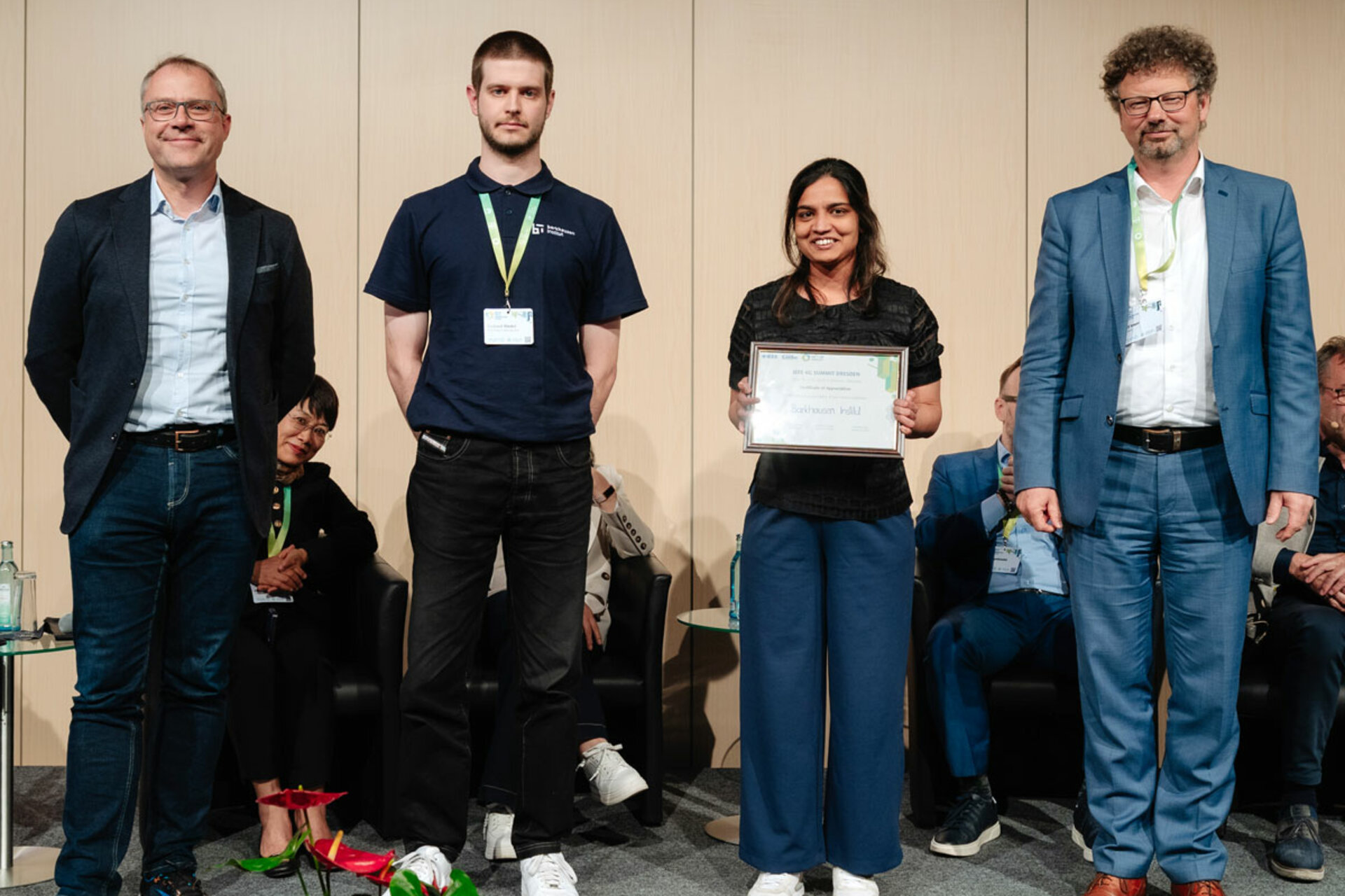 Vier Personen stehen nebeneinander, eine Frau hält eine Urkunde und lächelt, zwei Männer tragen Anzüge, ein Mann trägt ein schwarzes Polohemd mit Namensschild.