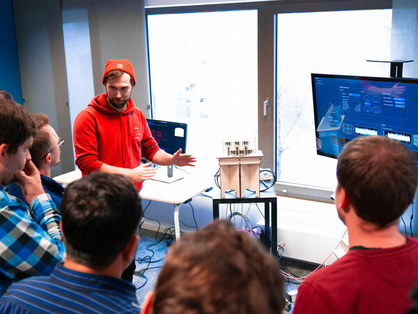 Ein Mann in einem roten Hoodie und einer roten Mütze erklärt einer Gruppe von Zuhörern etwas an einem technischen Modell. Im Hintergrund sind zwei Bildschirme mit Datenvisualisierungen zu sehen. Die Atmosphäre wirkt professionell und konzentriert.