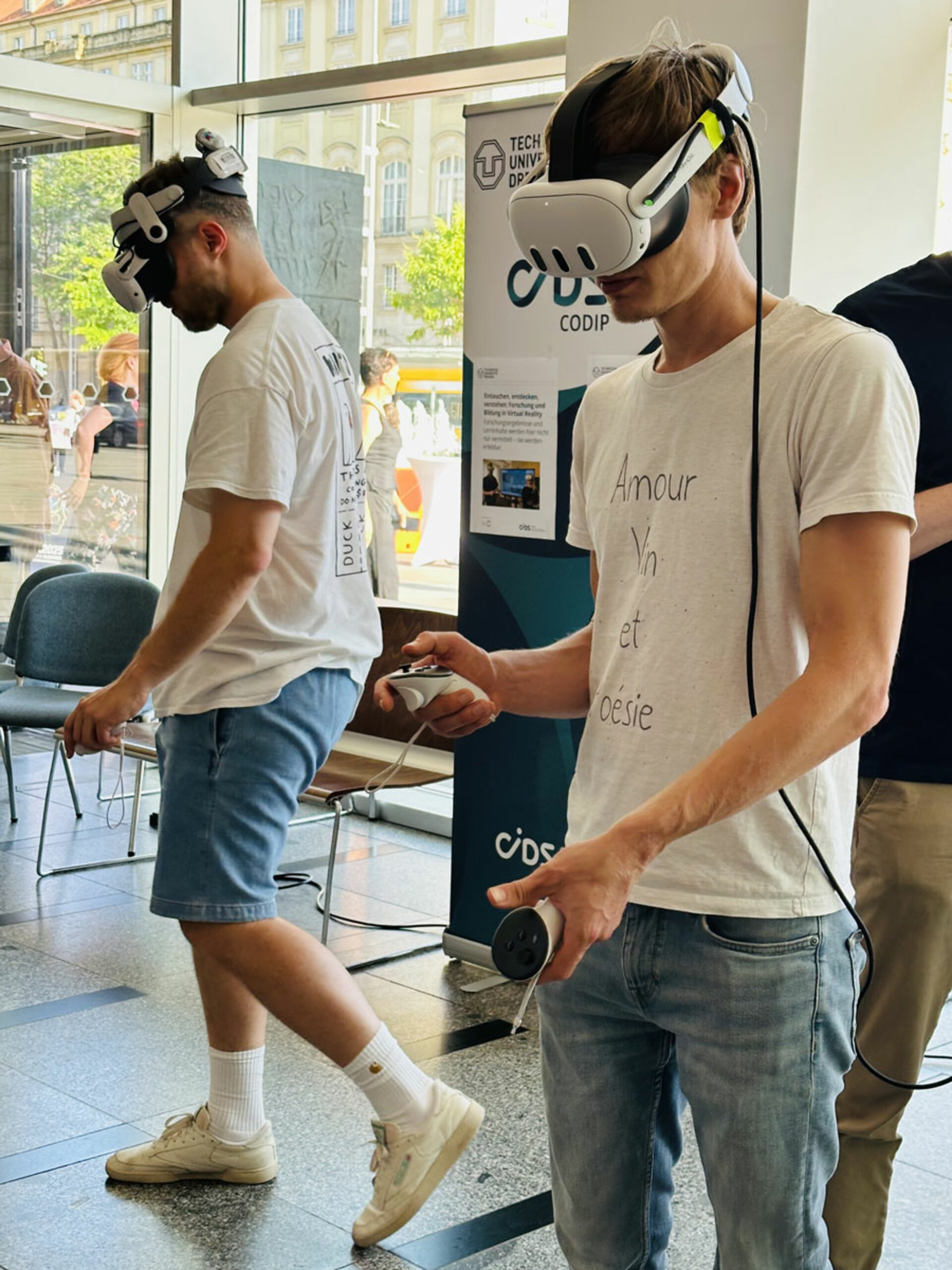 Zwei Personen, die VR-Headsets und legere Kleidung tragen, interagieren mit Virtual-Reality-Controllern in einem hellen Innenraum, konzentriert und vertieft in das Erlebnis.