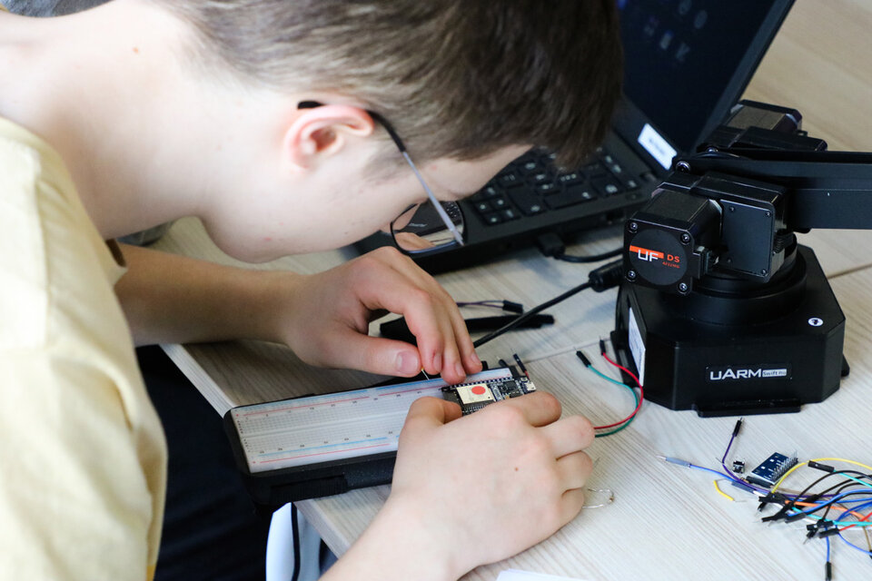 A young person is focused on assembling electronic components on a breadboard, with a robotic arm and a laptop nearby, conveying concentration and curiosity.