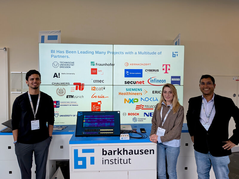 Three people standing behind a booth with a laptop and a sign reading 'barkhausen institut' and a screen behind showing logos of various partner organizations